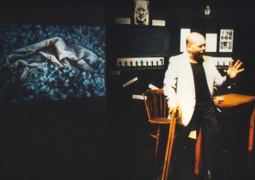 From the play Stalking Histories the character Mendel is sat on the edge of a table with his two walking sticks in one hand and he is gesturing with his other hand as he shouts at the audience. Behind him the image of the frosted child's jacket is showing on the screen. Performer - Garry Robson Photo - Felicity Shillingford 