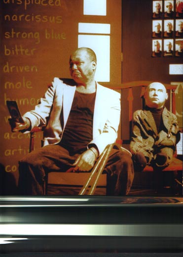 Production photo from Stalking Histories. The character Mendel is holding up a photograph. and scowling at it. Next to him on a love seat sits Dolly Boy, a smaller puppet version of Mendel. Behind them are words chalked on the wall - bitter, driven, mole. Mendel played by Garry Robson Photo by Felicity Shillingford